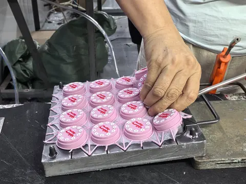 A bare hand lifting a freshly demoulded pink silicone part from a multi-cavity aluminium mould, runner still attached.